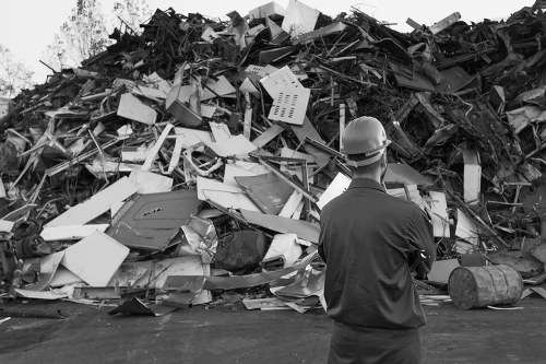 A man wearing a hard hat and dark work clothing stands on a paved surface, facing away from the camera, observing a large heap of mixed waste materials. The pile predominantly consists of disorganized electronic appliances, broken furniture, miscellaneous metal and plastic debris, and dismantled equipment, indicating a substantial clearance of bulky rubbish. The debris displays various textures, with some items appearing smooth and shiny, while others are rough or jagged, and their colours range from light grey and white to darker tones. The environment appears to be outdoors, possibly on a driveway or disposal site, with a background of scattered trees and an overcast sky. The scene illustrates the type of refuse collected during a comprehensive office or property clearance, typical of a professional rubbish removal service such as those provided by Office Clearance Thornton Heath, focused on removing accumulated waste efficiently from premises in the Thornton Heath area postcode. The overall atmosphere is neutral, with no visible activity beyond the man's assessment of the waste pile, emphasizing a methodical approach to rubbish clearance and environmental management.
