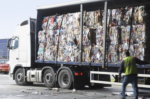 A large white commercial rubbish collection truck, with an open side compartment, is loaded with tightly packed bales of mixed recyclables, including paper, cardboard, plastic, and other waste materials, visible as compressed bundles with varying colors and textures. The truck is parked outdoors on a paved surface, with some scattered debris on the ground nearby. In the background, a person wearing a yellow high-visibility vest and dark clothing is walking past the truck, possibly a waste management worker. The scene appears to be part of a waste removal operation, consistent with commercial rubbish collection services in Thornton Heath or the surrounding area. The environment is bright, indicating daytime, and the overall setting emphasizes an organized, professional approach to waste disposal managed by companies like Office Clearance Thornton Heath.