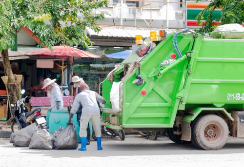 A bright green rubbish collection truck is positioned on a city street in Thornton Heath, with its rear compartment open, ready for waste collection. Two waste collection workers, dressed in grey uniforms with blue gloves and white hats, are actively engaged in handling black plastic refuse bags that are placed on the pavement in front of the truck. The workers are bending over, loading bags into the truck’s rear compartment, which contains various waste items. Behind the truck, there are small shops with colorful awnings, including a visible red-and-white striped canopy and another with a pink umbrella providing shade. A few pedestrians and parked scooters are also visible along the street, indicating an active urban environment. Surrounding the scene are green trees that add natural elements to the setting. The environment appears to be a mixture of commercial and residential area typical of Thornton Heath, with the waste collection process taking place on a clear day with natural daylight illuminating the scene. The presence of the Office Clearance Thornton Heath service is contextually relevant, emphasizing the local rubbish removal activity.