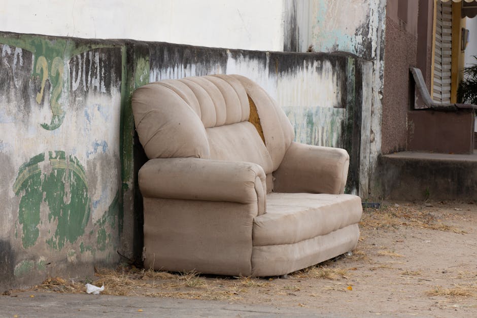 A burgundy leather sofa with visible creases and a smooth, slightly shiny texture is positioned upright, resting against a rough, dark stone wall on a paved outdoor surface. The sofa’s arms and back