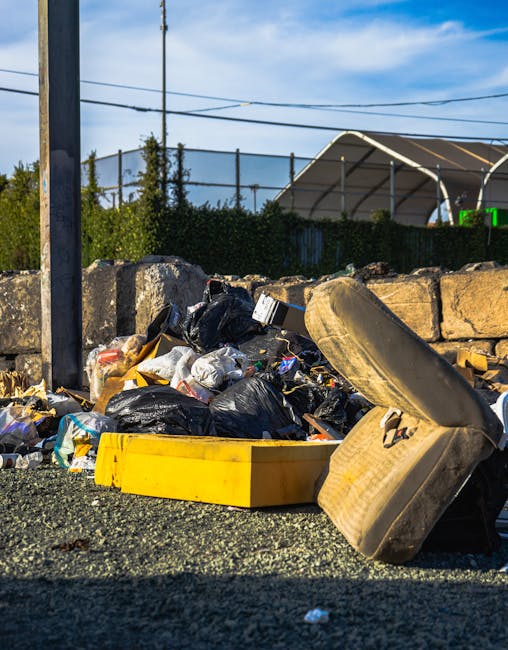 A large red metal waste skip with visible signs of rust and weathering is positioned against the corner of a modern building's dark green tiled wall and a plain grey concrete wall. The skip is partial