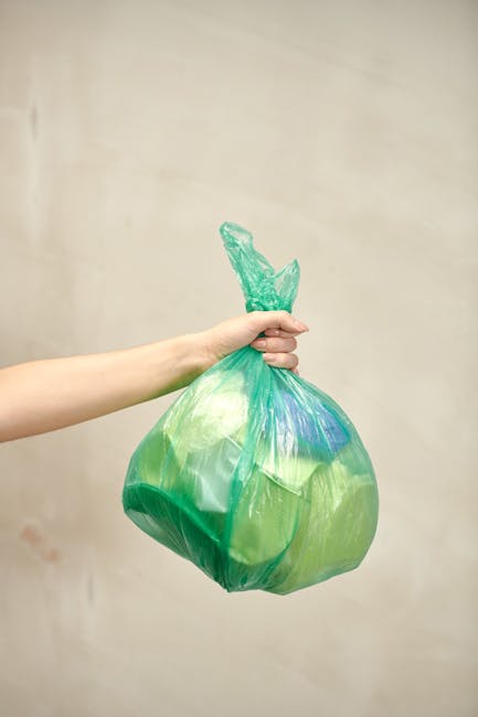 A middle-aged man with dark, slightly wavy hair, wearing a black T-shirt with white graphic print on the front, is seen disposing of rubbish into a tall, stainless steel public trash bin located on a 
