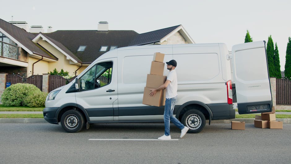 A man dressed in casual clothing, including a white t-shirt, blue jeans, white sneakers, and a black cap, is standing next to a white commercial van parked on a residential street. He is holding three