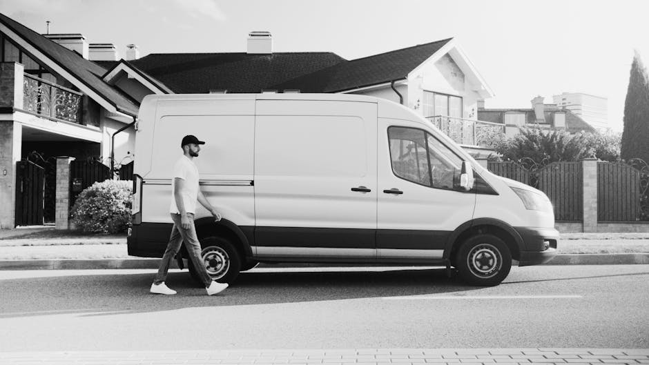 A man dressed in casual clothing, including a white t-shirt, blue jeans, white sneakers, and a black cap, is standing next to a white commercial van parked on a residential street. He is holding three