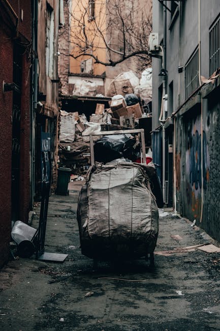A narrow urban alleyway filled with a large pile of garbage bags, cardboard boxes, and miscellaneous waste materials stacked against a backdrop of old brick and concrete buildings. In the foreground, 