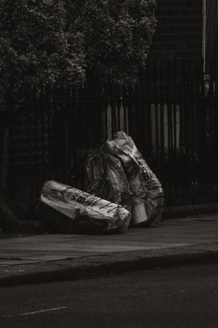 A narrow urban alleyway filled with a large pile of garbage bags, cardboard boxes, and miscellaneous waste materials stacked against a backdrop of old brick and concrete buildings. In the foreground, 